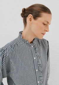 Vrouw met een knot in haar haar, gekleed in een zwart-wit geblokte blouse met een gerimpelde kraag, kijkt omlaag tegen een effen achtergrond.