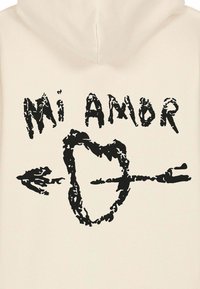 Felpa crema con stampa grafica nera contenente il testo "Mi Amor" sopra un design di cuore e freccia. Tessuto morbido, vestibilità regolare.