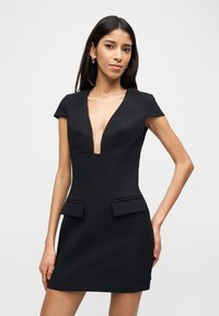 Γυναίκα που φοράει εφαρμοστό μαύρο φόρεμα με βαθύ V ντεκολτέ και κοντά μανίκια, στέκεται μπροστά σε λευκό απλό φόντο.