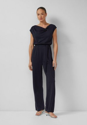 Frau steht vor einer schlichten Wand und trägt einen ärmellosen marineblauen Overall mit tailliertem Gürtel und silbernen offenen Absatzschuhen.