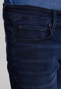 Tmavě modré džíny z denimu s hladkou texturou, standardním designem s pěti kapsami, kovovými detaily a jemným vyblednutím na nohavicích.