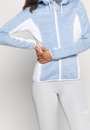 Kobieta w jasnoniebieskiej i białej kurtce z kapturem na zamek, ze skrzyżowanymi rękami na wysokości talii, ubrana w dopasowane jasnoszare spodnie na jednolitym tle.