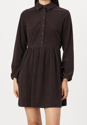 Kobieta w ciemnobrązowej, sztruksowej sukience z długim rękawem, zapinanej na guziki z przodu, z marszczoną talią, pokazana od ramion do połowy uda.