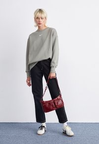 Person med blont hår som bär en grå Nike-tröja, svarta byxor, vita sneakers och håller en röd läderhandväska, står på en blå matta mot en vit vägg.