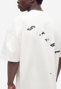 Άτομο που φοράει ένα λευκό φαρδύ t-shirt με τη λέξη "B-S-A-C-U-E" τυπωμένη διαγώνια στην πλάτη.
