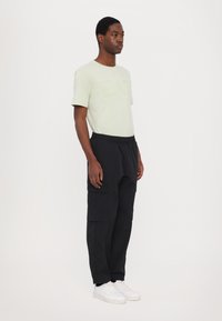 T-shirt vert clair à manches courtes avec logo en relief, associé à un pantalon cargo noir ample avec des poches latérales, porté avec des baskets blanches.