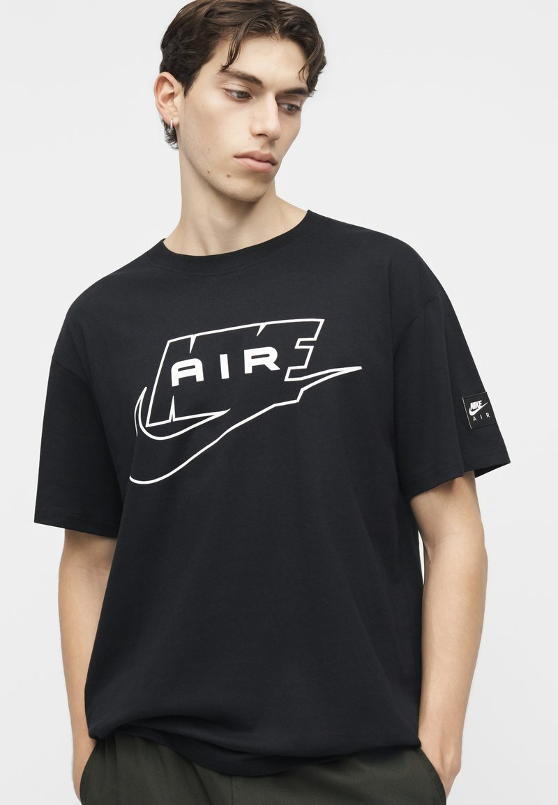 Junger Mann mit braunen Haaren trägt ein schwarzes Nike Air T-Shirt mit weißem Logo, Hände in den Taschen, schaut nach unten und zur Seite.