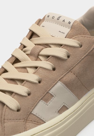 Αθλητικό παπούτσι από σουέτ σε μπεζ χρώμα με κρεμ κορδόνια, λευκό λογότυπο "H" στο πλάι και ετικέτα "HOGAN MILANO" στη γλώσσα.