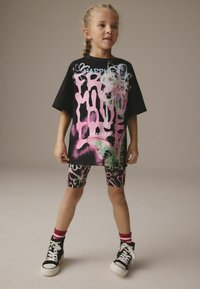 T-shirt noire surdimensionnée avec des graphiques colorés de style graffiti, associée à un short à motifs et des baskets montantes noires avec des semelles blanches.
