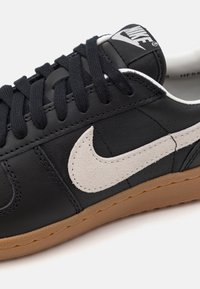 Baskets Nike en cuir noir avec swoosh en daim blanc, lacets noirs et semelle en gomme, présentées en vue rapprochée de côté.