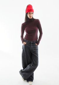 Bordeauxfarbenes Langarmshirt kombiniert mit lockeren, dunkelgrauen Jeans. Eine leuchtend rote gerippte Mütze mit einem kleinen schwarzen Bandakzent. Weiße Schuhe.