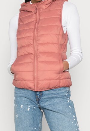 Chaleco acolchado sin mangas en rosa suave con capucha y cremallera delantera. Presenta costuras horizontales con bolsillos laterales, usado sobre una camiseta de manga larga blanca.