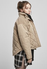 Beige Steppjacke mit einem gepolsterten Design und weichem, beigefarbenem Kragen. Hat einen verkürzten, übergroßen Schnitt und eine geknöpfte Vorderseite. Getragen über einem karierter Rock.