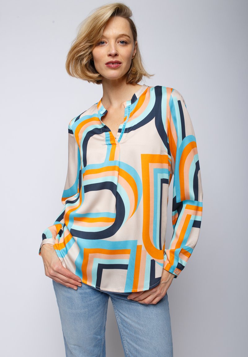 Langsleeve blouse in lichtbeige met gedurfde geometrische patronen in blauw, oranje en marineblauw. Heeft een V-hals en een ontspannen pasvorm. Gladde stof.