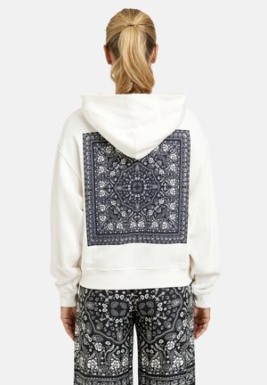 Frau mit blondem Pferdeschwanz, die einen weißen Kapuzenpullover mit großem quadratischem schwarz-weißem Paisley-Muster auf dem Rücken und passende gemusterte Hosen trägt.