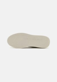 Suela de tenis beige claro con patrones texturizados, que presenta ranuras y un logo en relieve en el talón. Diseño plano, material de goma.