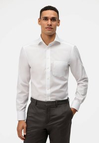 Camicia bianca a maniche lunghe con colletto button-down e tasca sul petto, caratterizzata da una consistenza liscia e una vestibilità classica. Abbinata a pantaloni scuri.