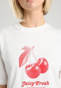 Camiseta de algodón blanca con un gráfico rojo de dos cerezas y una hoja, con el texto "Jugoso Fresco" en rojo debajo del diseño.