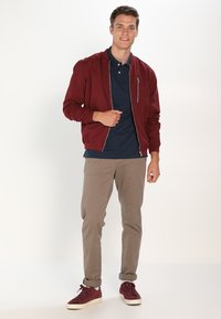 Jachetă bomber maro cu fermoar în față, cămașă polo bleumarin, pantaloni bej și adidași burgundy. Textură netedă și design ajustat.