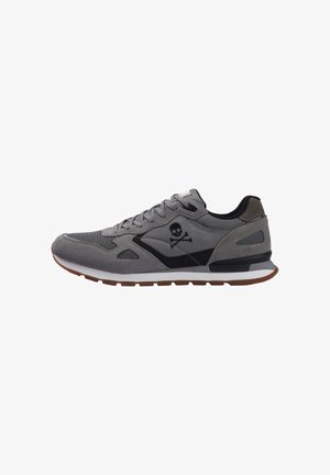 Zapatilla deportiva gris con parte superior de ante, detalles de malla, elementos en negro y un gráfico de calavera y huesos cruzados. Entresuela blanca y suela de goma.