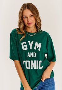 Zelené bavlnené tričko s veľkým bielym grafickým textom "GYM AND TONIC." Krátke rukávy a voľný strih. V kombinácii s modrými denimovými džínsami.