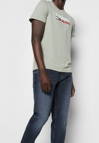 Ljusgrön bomullströja med "TOMMY JEANS"-logotyp i röd och svart, kombinerad med mörkblå denimjeans. Enkel design med korta ärmar.