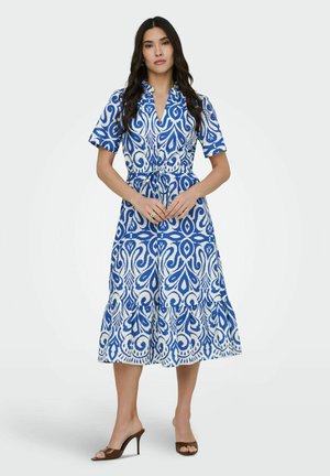 Ženska z dolgimi temnimi lasmi, oblečena v modro-belo vzorčasto midi obleko s kratkimi rokavi in rjavimi sandali z visoko peto.