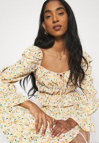 Vestido floral com mangas bufantes, apresentando decote quadrado, frente com cordão de ajuste, base em amarelo pastel e padrões de flores verdes e laranja.