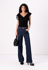 Svart rynket topp med volangermar, kombinert med brede mørke denimjeans. Modellen holder en svart håndveske og har på seg spisse hæler.