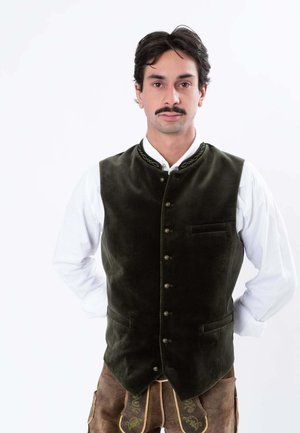 Man with dark hair and mustache wearing a dark green buttoned vest over a white shirt and brown traditional embroidered pants, standing with hands behind back.