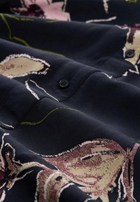 Dunkelblauer Stoff mit abstraktem Blumenmuster in Beige, Lila und Grün, versehen mit einem Knopf und sichtbaren Nähten.