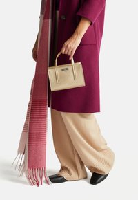 Frau in beigen weit geschnittenen Hosen, schwarzen Schuhen und einem magentafarbenen Mantel, die eine kleine goldene Handtasche und einen pink-roten Fransen-Schal hält.