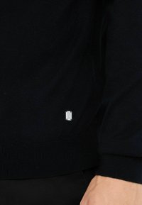 Maglione blu navy realizzato in materiale lavorato a maglia, con orlo e polsini a coste. Include una piccola etichetta in logo argentato sul lato sinistro in basso.