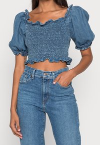 Top court en denim avec des manches bouffantes, texture smockée et encolure froncée. Associé à un jean en denim taille haute de la même teinte.
