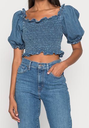 Denim crop top dengan lengan puff, tekstur smocked, dan leher ruffle. Dipadukan dengan celana jins high-waisted dalam warna senada.