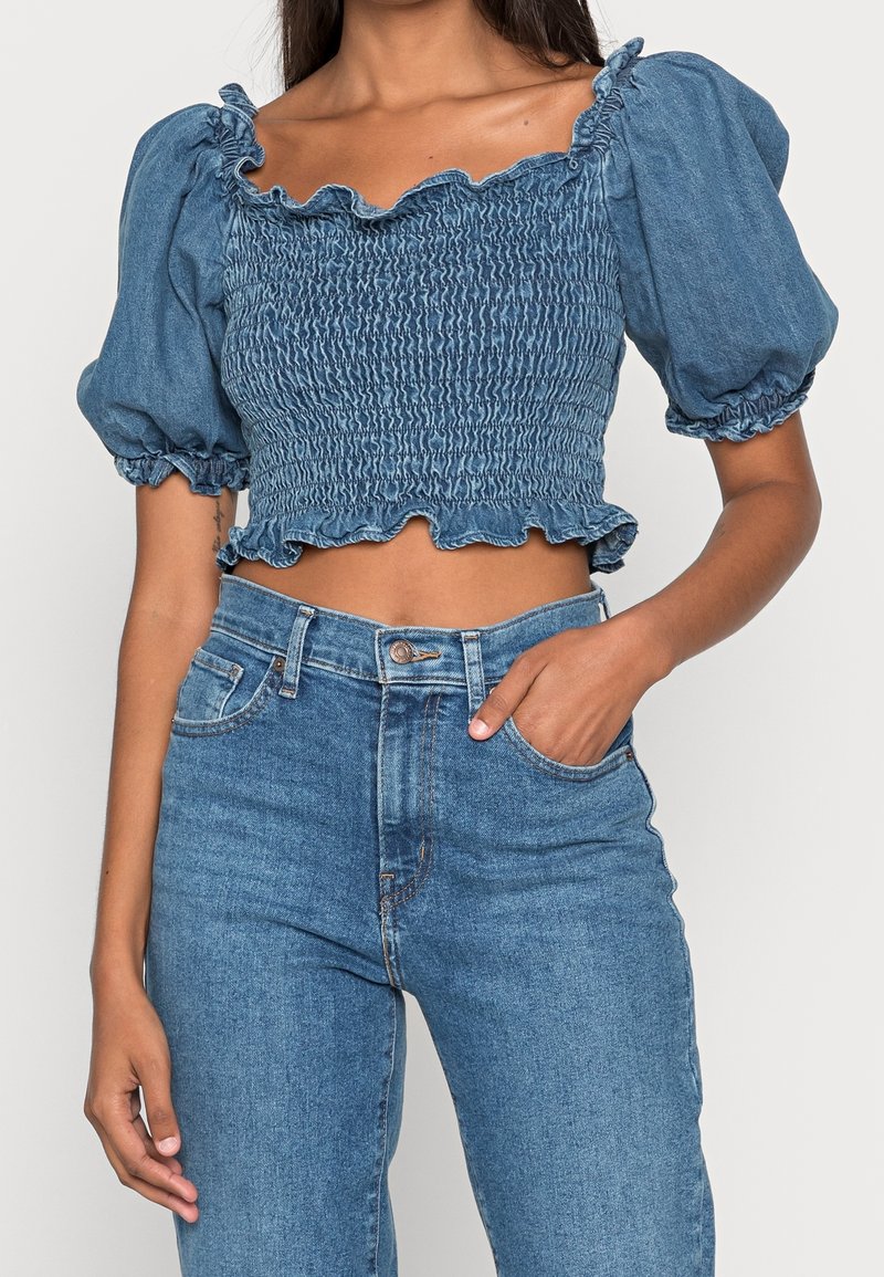 Top court en denim avec des manches bouffantes, texture smockée et encolure froncée. Associé à un jean en denim taille haute de la même teinte.