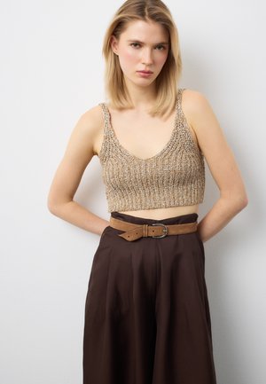 Donna con capelli biondi che indossa un top corto di maglia beige e pantaloni a vita alta marrone scuro con una cintura in pelle scamosciata color cognac.
