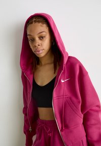 Magenta mikina na zips s veľkou kapucňou, predným vreckom a malým bielym logom Nike. Nosená cez čierny tielko a zodpovedajúce nohavice.