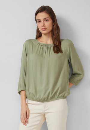 Jeune femme aux longs cheveux bruns portant un chemisier vert clair à manches trois-quarts et un pantalon crème, debout devant un mur gris uni.