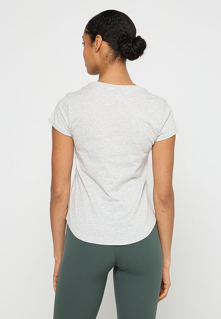 Frau mit dunklem Haar im Dutt trägt ein hellgraues Kurzarmshirt und dunkle grüne Leggings und steht mit dem Rücken zu einem schlichten weißen Hintergrund.