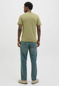 Olijfgroen T-shirt met korte mouwen, gecombineerd met lichtblauwe rechte jeans en beige schoenen. De stof lijkt zacht en casual.