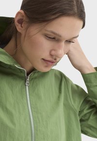 Veste légère verte avec fermeture éclair sur le devant, col montant et texture lisse. Dotée d'une fermeture éclair argentée et d'une coupe décontractée.