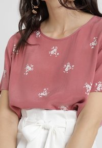 Pink kortærmet bluse med hvidt blomsterprint, rund halsudskæring og let stof. Bæres med en hvid høj talje nederdel, der er bundet.