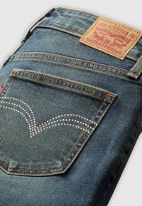 Poche arrière d'un jean en denim bleu avec des coutures orange, un design en strass et une étiquette en cuir Levi Strauss & Co. de couleur beige au-dessus.