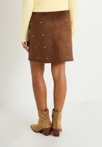 Falda de ante marrón con ojales plateados, hasta la rodilla, con cremallera oculta. Combinada con botas de tobillo beige con tacón cuadrado.