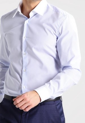 Man draagt een lichtblauw overhemd met fijne strepen en knoopmanchetten, en marineblauwe broek, terwijl hij zijn overhemd ter hoogte van de taille recht trekt.