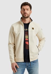 Veste beige clair avec une fermeture éclair à l'avant, un col noir et des panneaux d'épaule texturés. Comprend des poches latérales et un petit logo sur la poitrine.