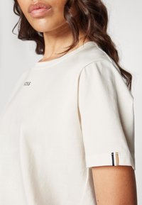 Cream-colored cotton T-shirt with a round neckline, short sleeves, and a small logo on the chest; includes a striped accent on the sleeve hem.
