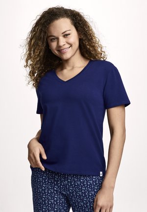 Frau mit lockigem Haar lächelt und trägt ein marineblaues V-Ausschnitt-T-Shirt sowie dunkelblaue, gemusterte Hosen, posiert mit einer Hand in der Tasche.