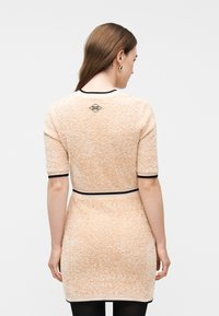 Γυναίκα με καστανά ίσια μαλλιά, φορώντας μια υφασμάτινη μπεζ κοντομάνικη φορέμα με μαύρο περίγραμμα, εμφανιζόμενη από την πίσω πλευρά μπροστά σε απλό φόντο.